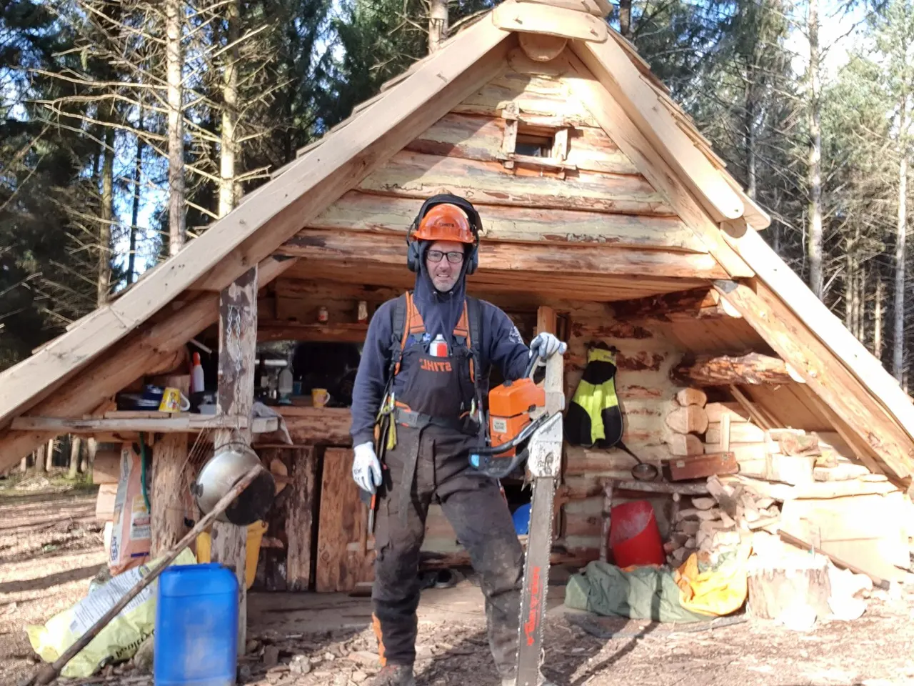 Person wearing safety helmet and protective gear holding chainsaw outside rustic wooden lodge surrounded by conifer trees.