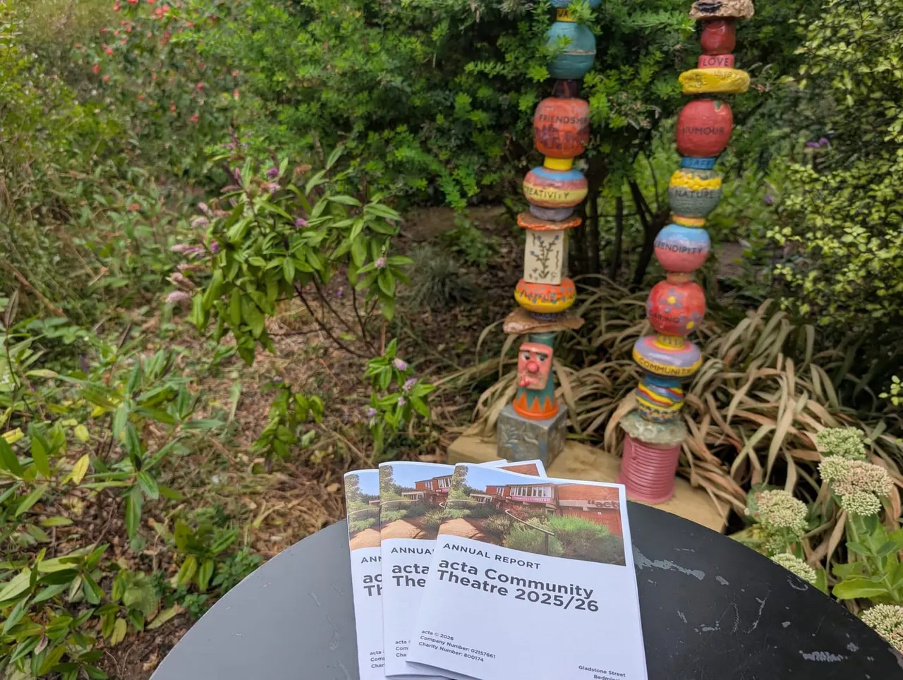 Colourful stacked garden sculptures and plants beside an annual report for acta Community Theatre on a garden table.