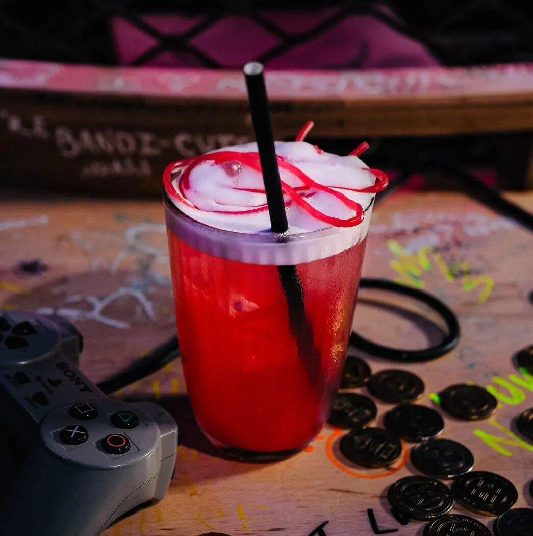 Red cocktail with ice and red ribbon garnish on gaming table with arcade controls nearby.