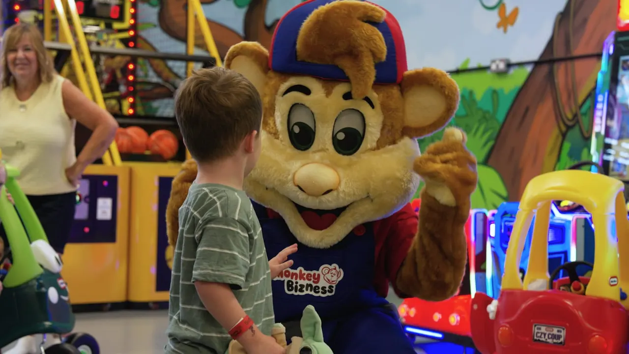 Young child meeting a costumed monkey mascot character in an indoor soft play arcade with illuminated games and rides.