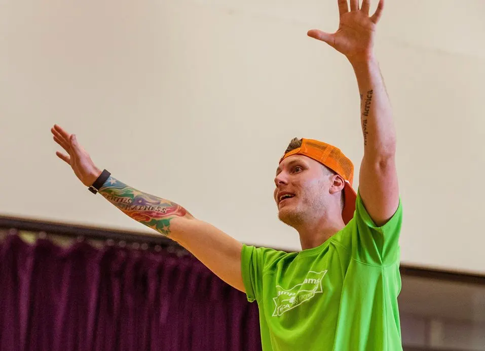 Man in bright green shirt and orange headband performing or presenting with arms raised theatrically indoors.