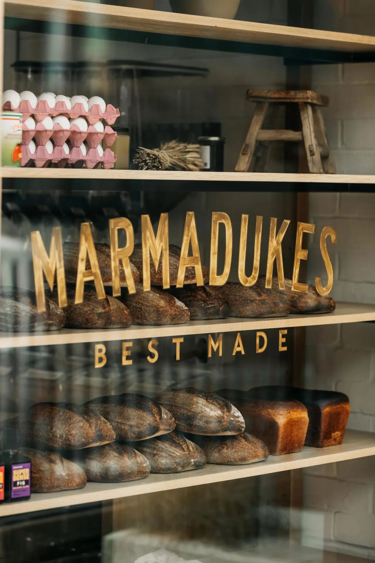 Display cabinet with freshly baked bread loaves and egg cartons, featuring gold lettering reading "Marmadukes Best Made".