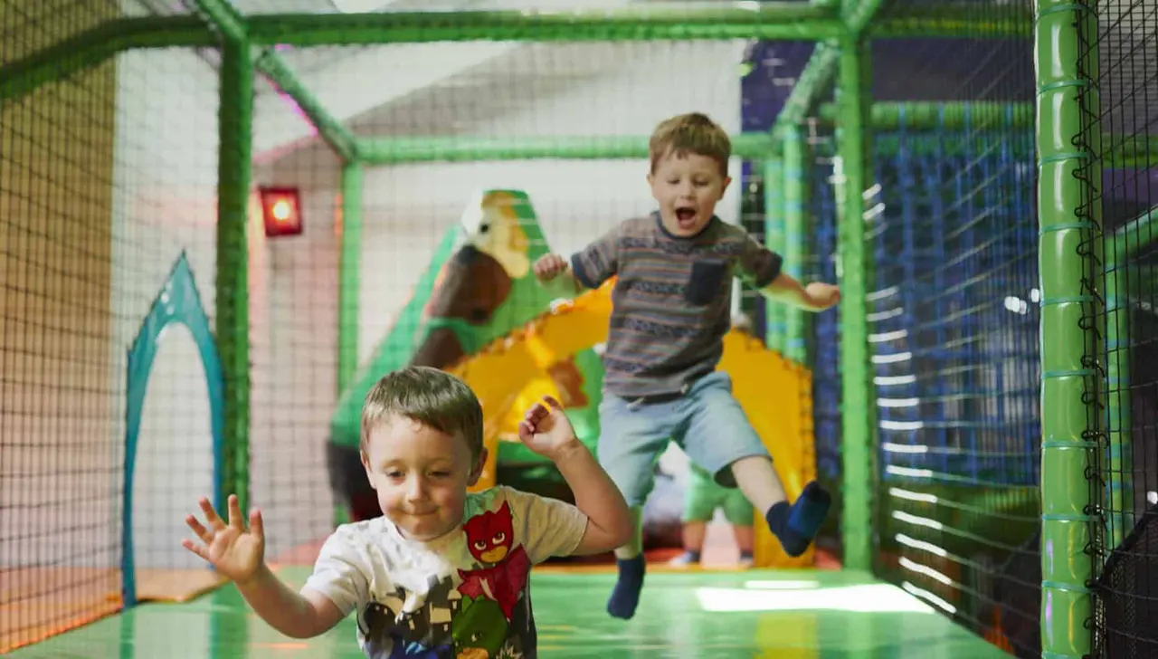 Young children playing in a colourful indoor soft play maze with green netting and foam obstacles.