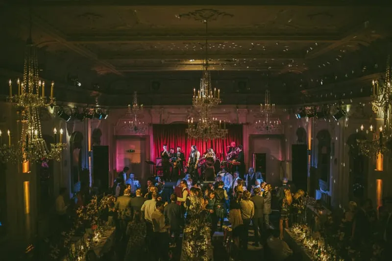 A crowd of people dancing and socialising in an ornate ballroom with a live band performing on stage under chandeliers and pink lighting.