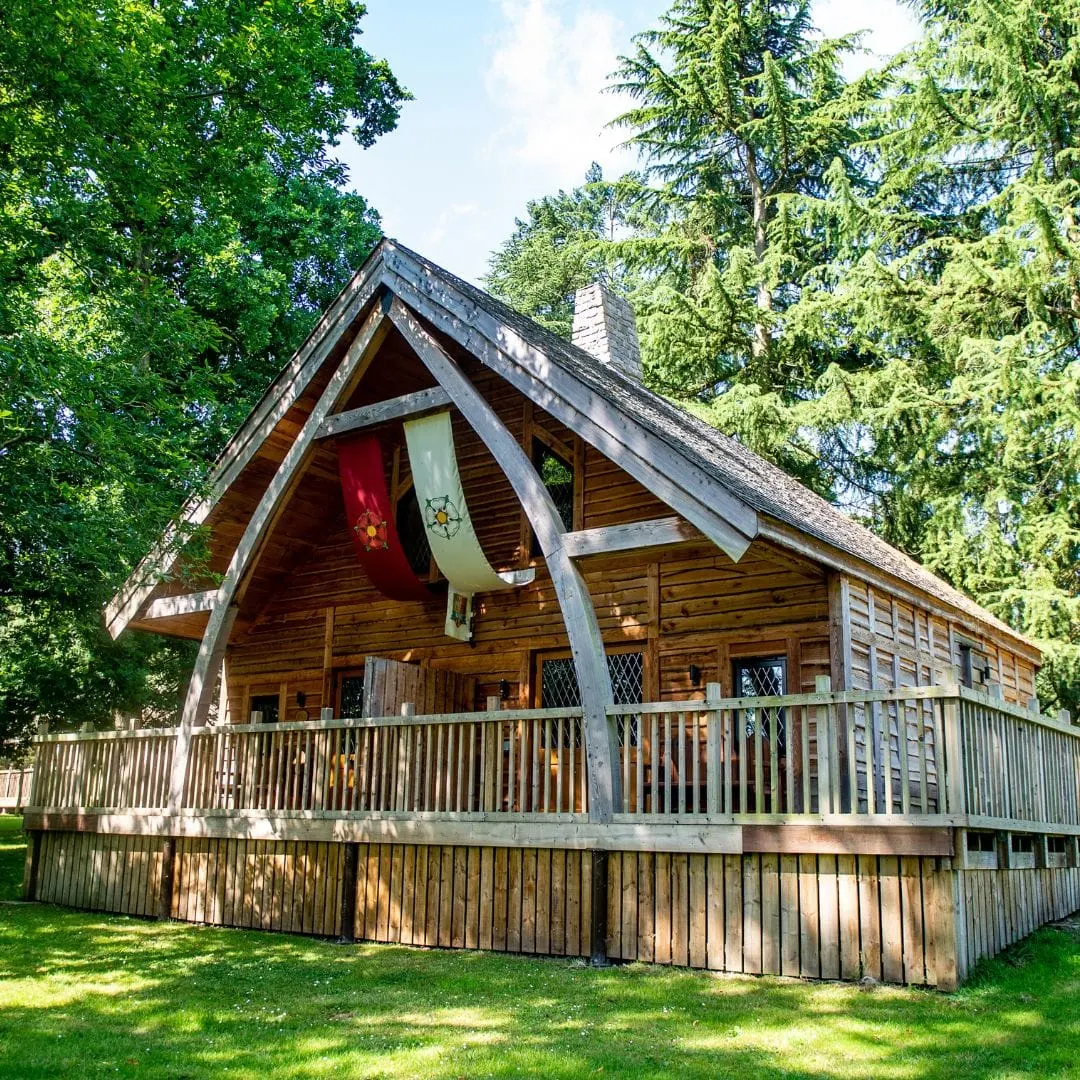 A timber-framed medieval-style wooden cabin with a steep pitched roof and large arch, surrounded by tall trees and green grass.