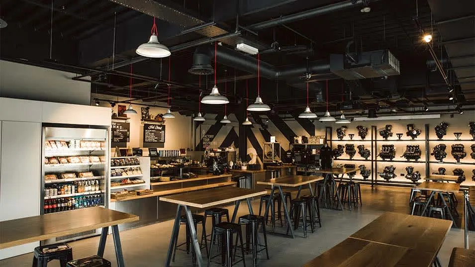Industrial-style tasting room with high ceilings, pendant lights, bar seating, and display shelves of merchandise.