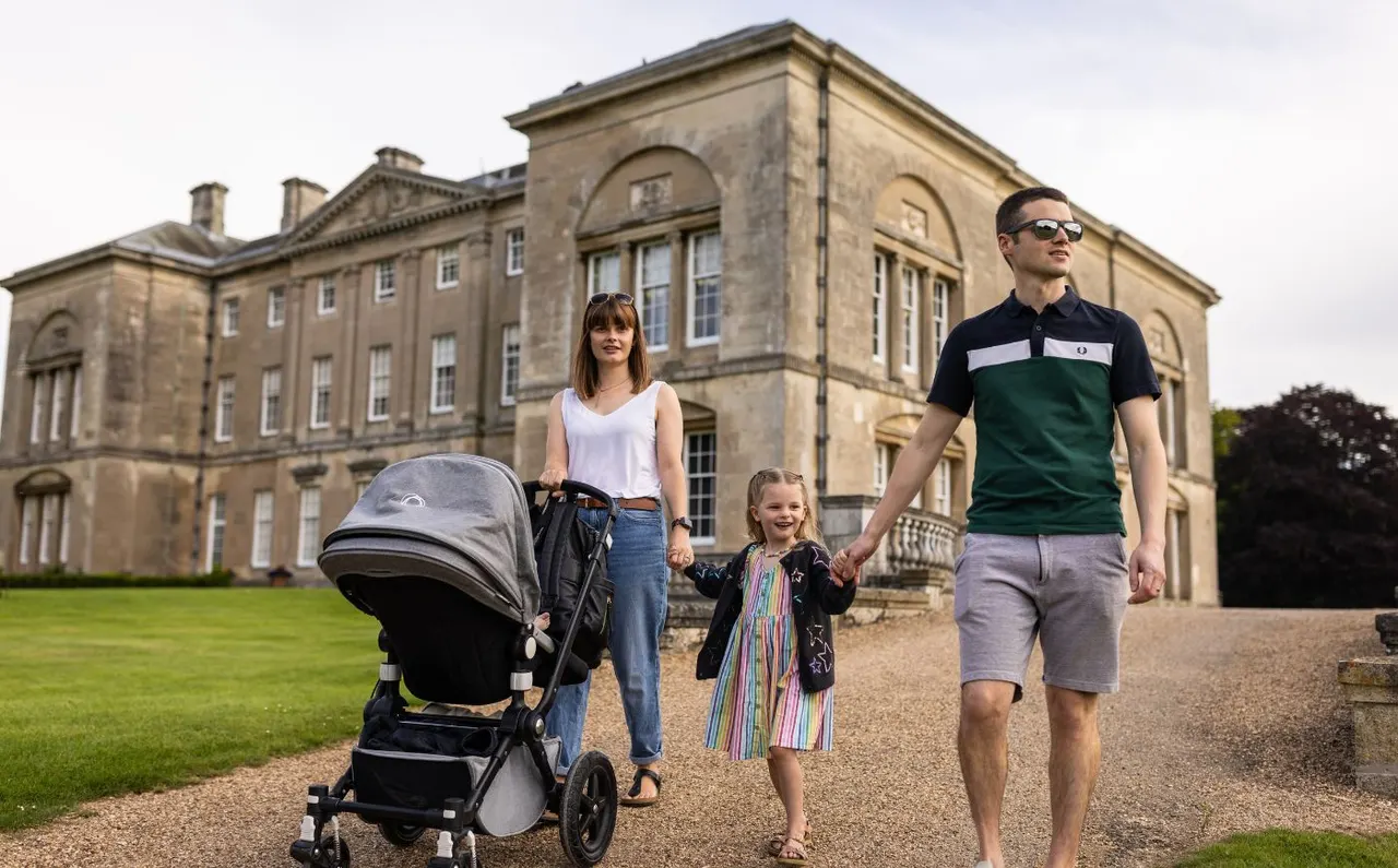 Family walking on a gravel path in front of a large stone stately home, pushing a pram.