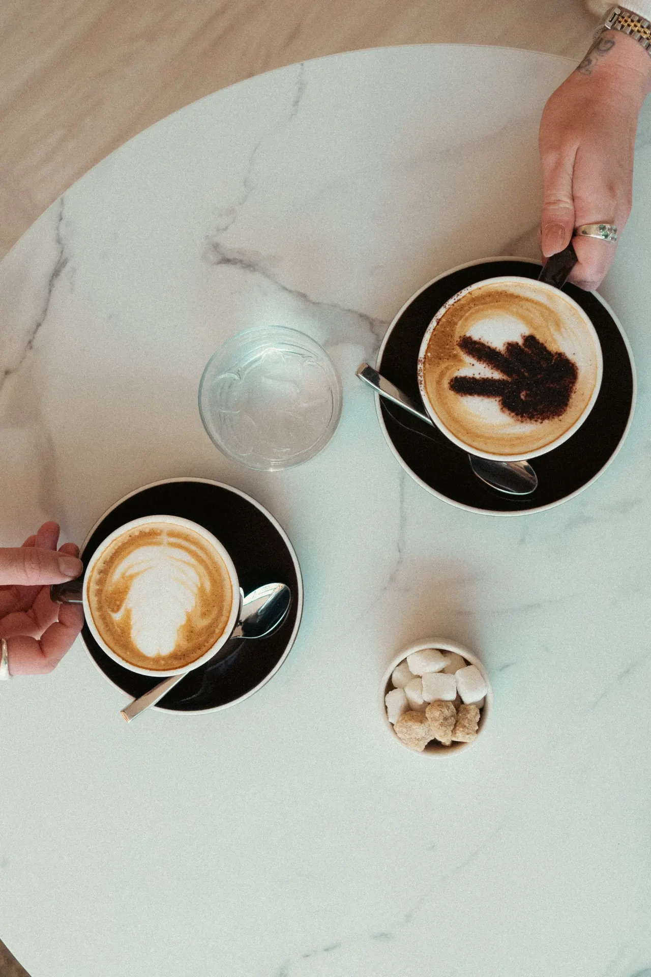 Two people holding cappuccinos with latte art on a marble table with water and sugar cubes.