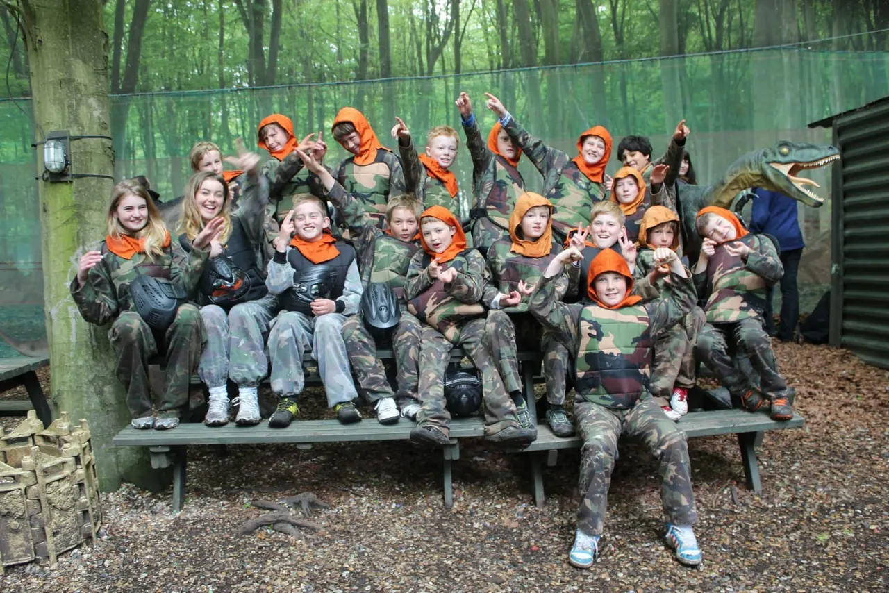 Group of children in camouflage gear and orange caps posing together on a bench in a woodland setting.