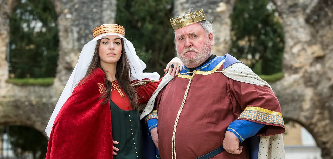 Two people in medieval royal costume pose together against stone ruins and greenery.