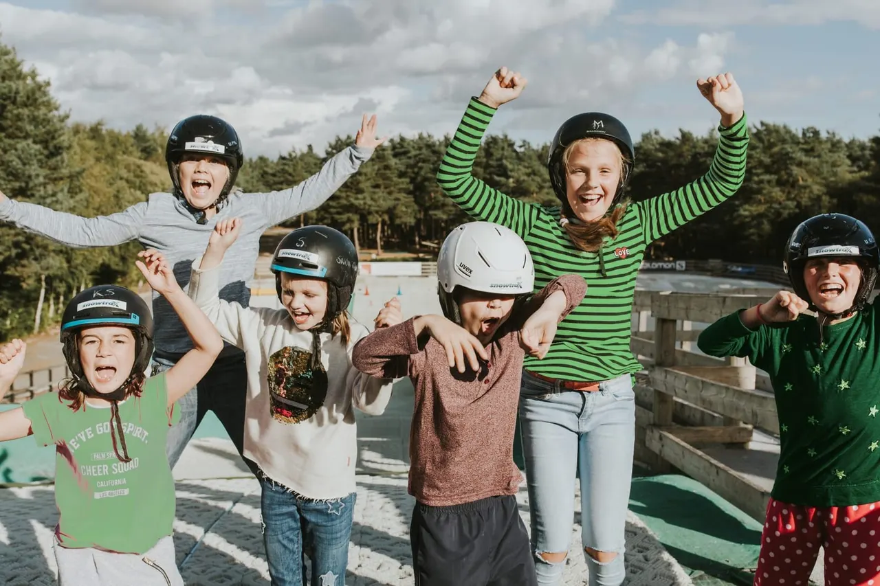 Group of children and a young instructor wearing helmets celebrating outdoors at a snow sports venue, with forest and buildings in background.