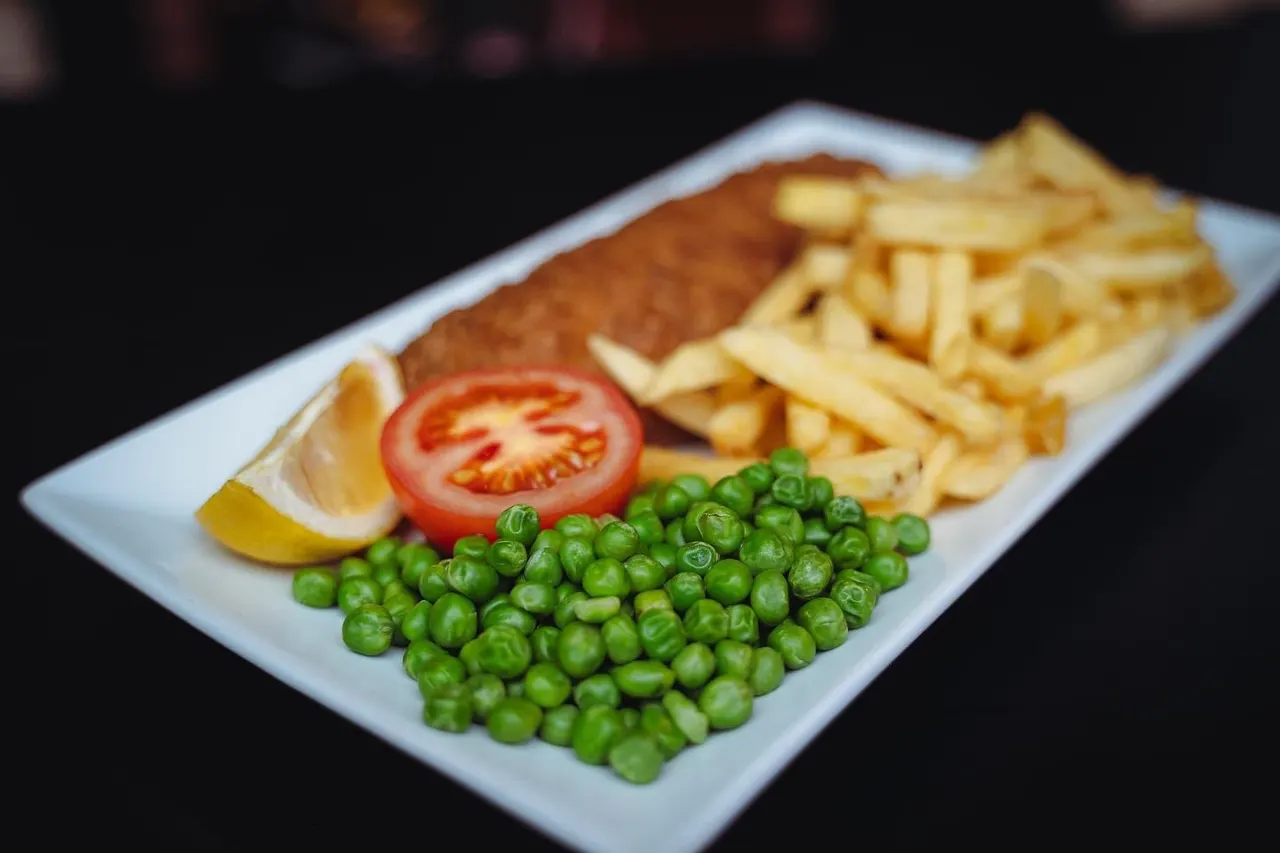 Breaded fish fillet with chips, peas, tomato slice and lemon wedge on a white plate.