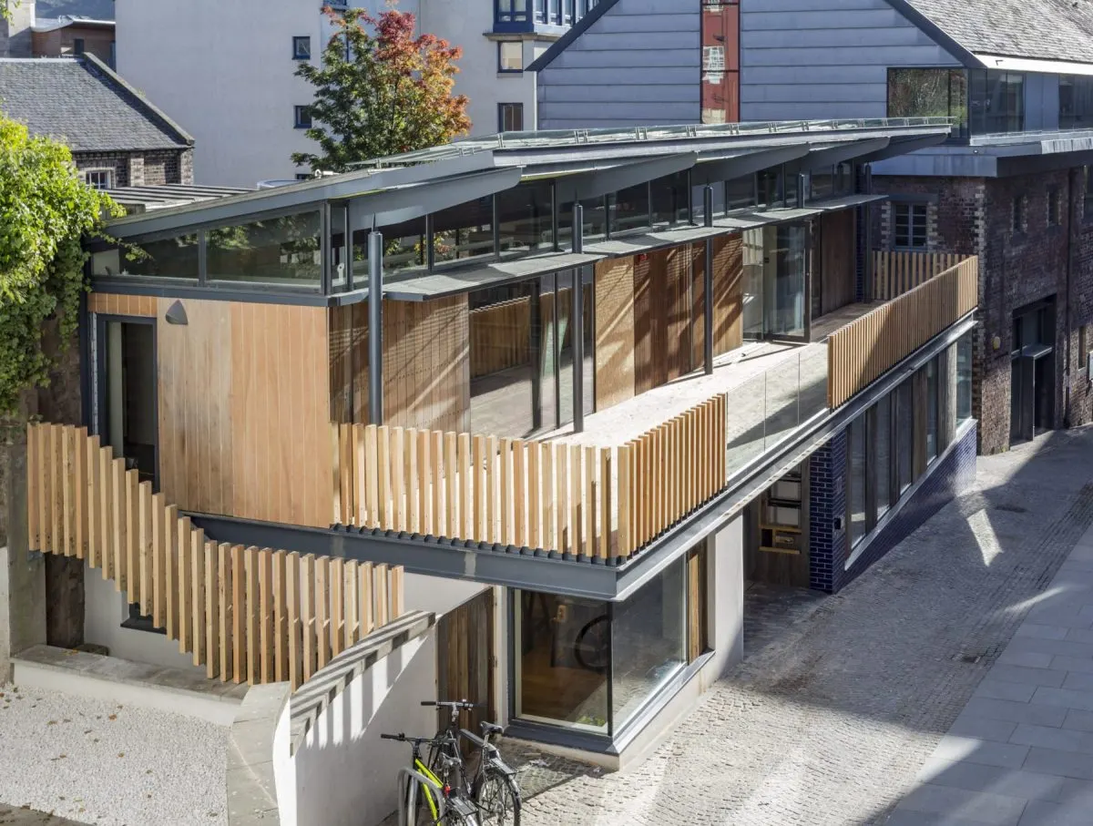 Modern timber and glass building with wooden railings beside a courtyard, adjacent to historic stone buildings.