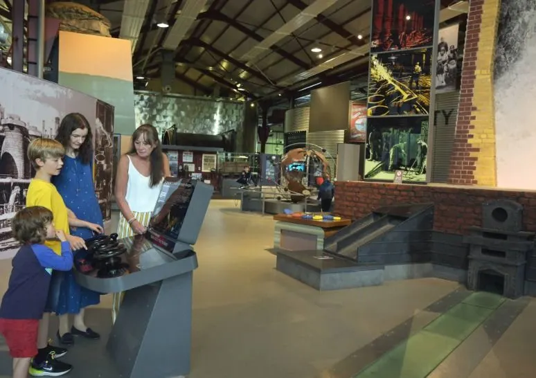 Adult and two children exploring interactive displays in an industrial heritage museum with exposed brick walls and period machinery.