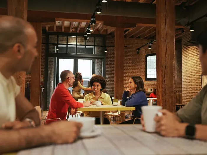 Adults socialising over drinks in a brick-walled café space with wooden beams and large windows.
