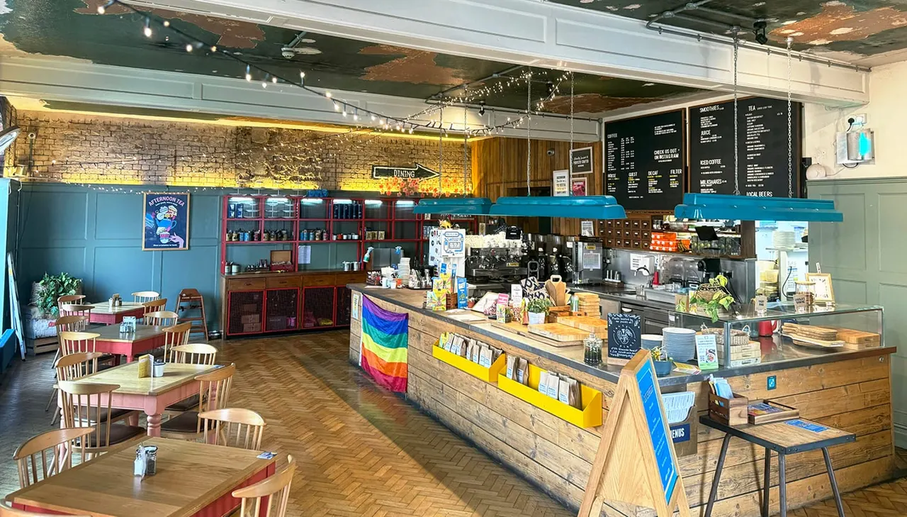 Interior of a modern café with wooden counter service area, dining tables, blue and red vintage furniture, and rainbow pride flag decoration.