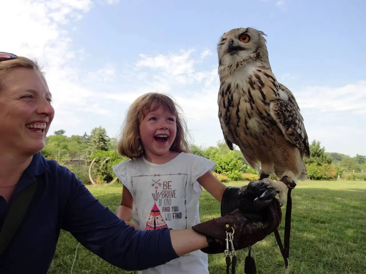 An adult and child hold a large owl perched on a gloved arm outdoors, both smiling at the bird.