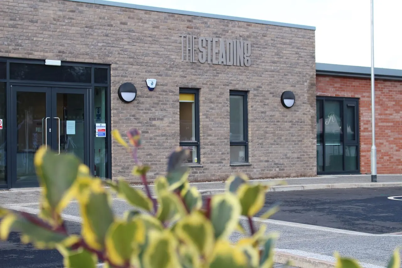 Modern brick building entrance with "The Steading" signage, black-framed doors and windows, with succulent plants in foreground.