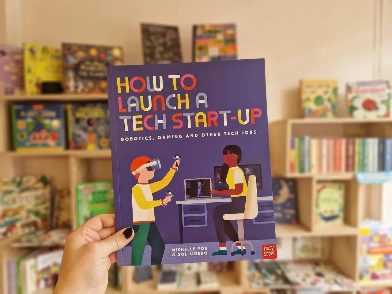 Hand holding a children's book titled "How to Launch a Tech Start-Up" in front of shelves of books in a bookshop.