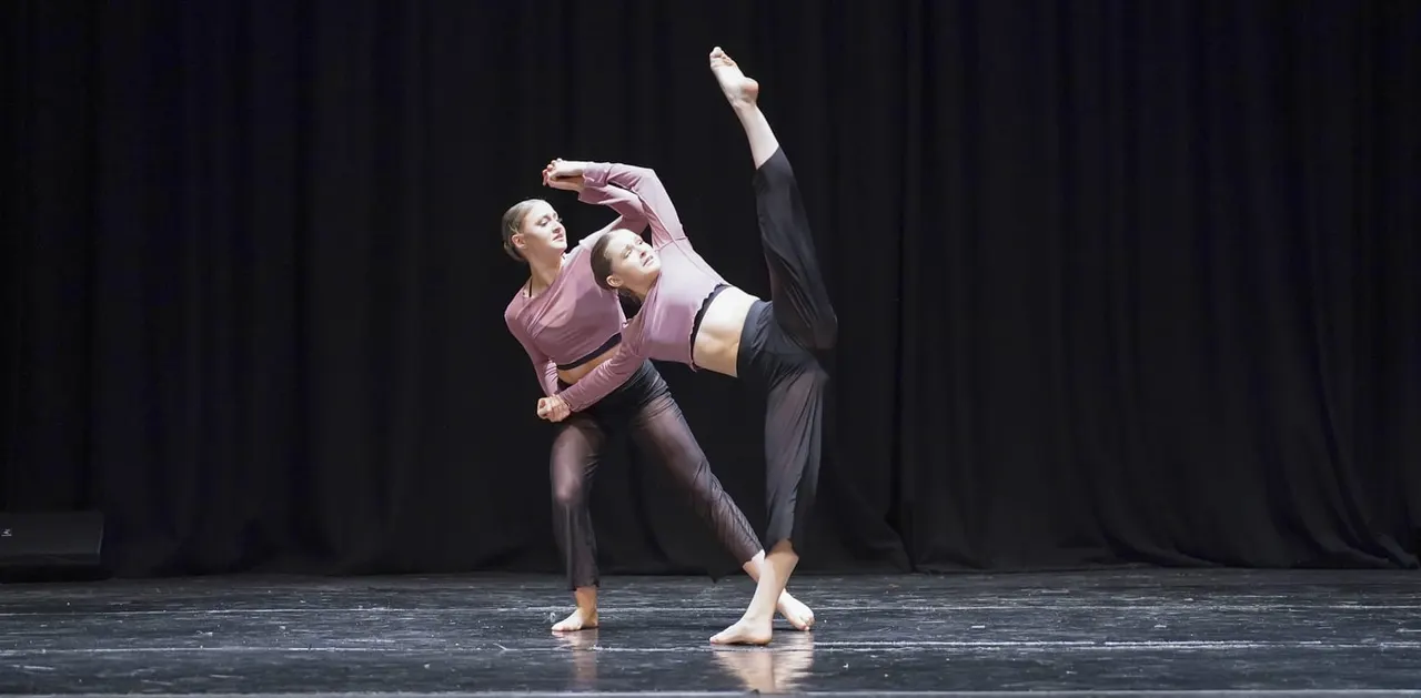 Two dancers performing a contemporary dance piece on stage, one lifting the other with an extended leg.