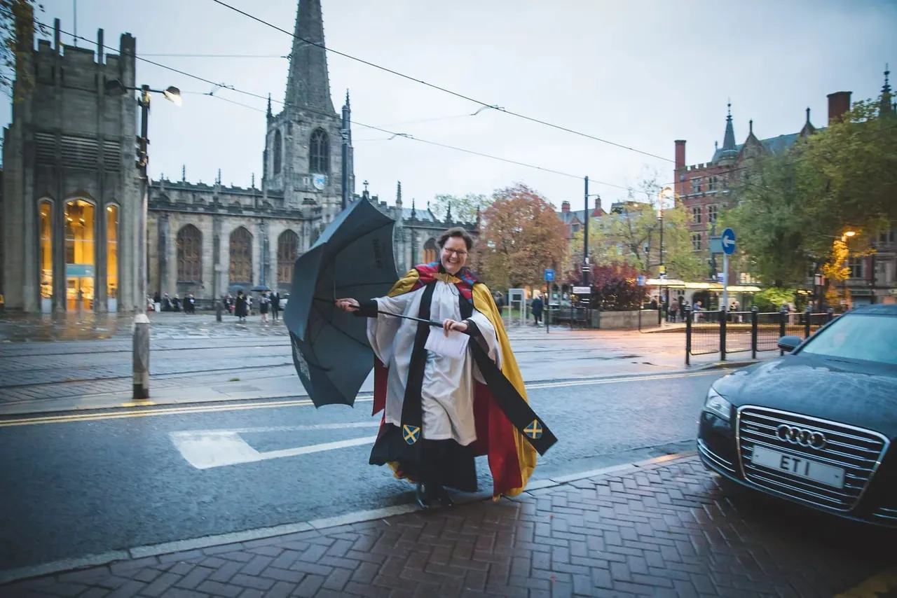 Person in academic robes holding an umbrella in a city square with a Gothic cathedral and historic buildings in the background.