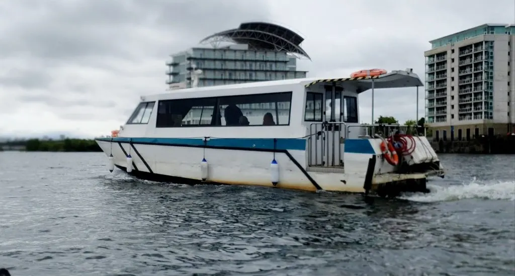 White and blue passenger ferry moored in shallow water near modern waterfront buildings.