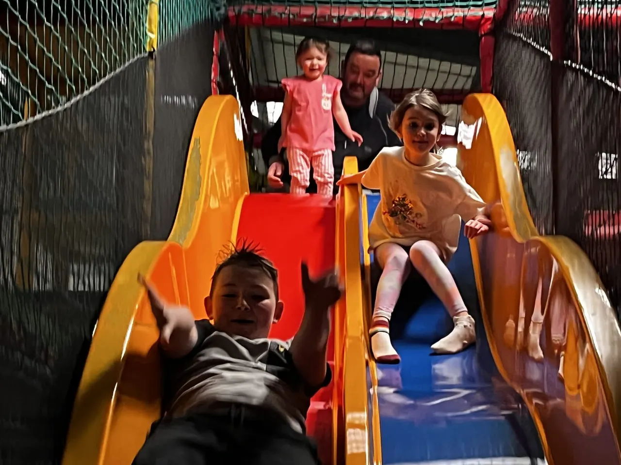 Children and an adult enjoying a bright yellow and red indoor slide at a play centre.