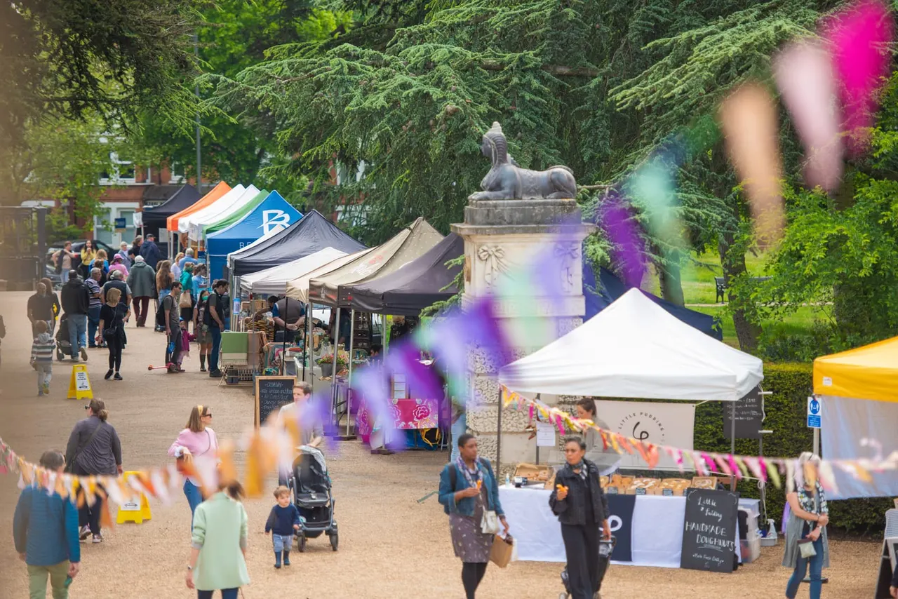 Outdoor market or festival with visitors browsing market stalls and tents surrounded by historic grounds and mature trees.
