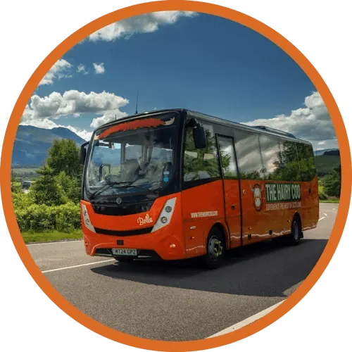 An orange tour bus parked on a road with mountains and sky in the background.