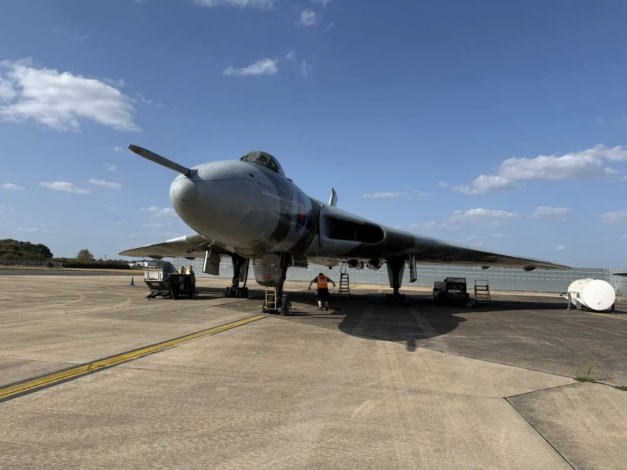 A vintage military aircraft on an airfield with ground staff and service vehicles beneath a clear blue sky.