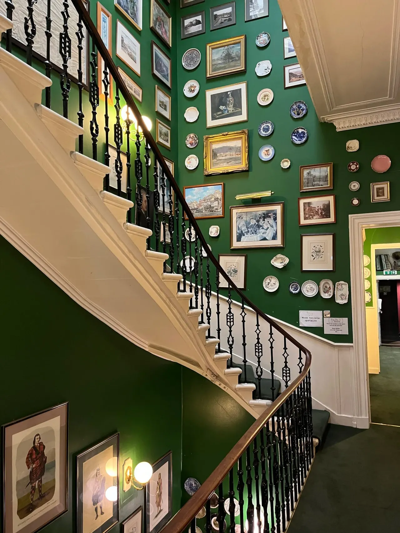Interior staircase with green painted walls displaying framed prints, plates and ceramics arranged gallery-style.