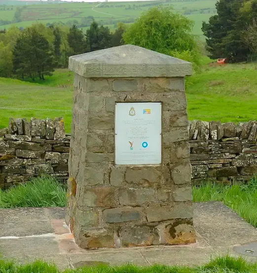 Stone monument with information plaque in rural green landscape bordered by dry stone walls.