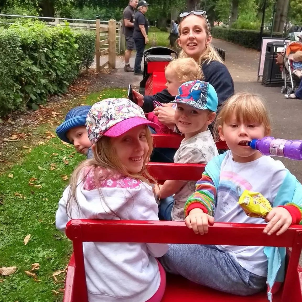 Woman and four young children riding together in a red open-air train car on a leafy park pathway.