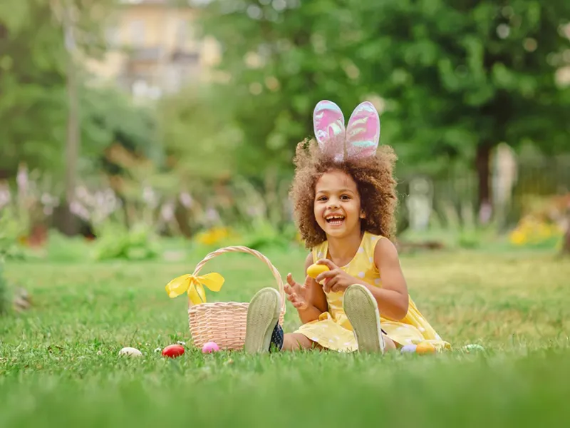Child wearing bunny ears and yellow dress sitting on grass with Easter basket and coloured eggs.