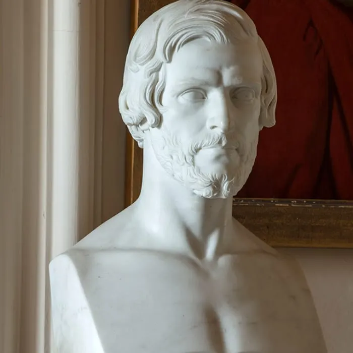 White marble bust sculpture of a bearded man displayed indoors against a burgundy wall.
