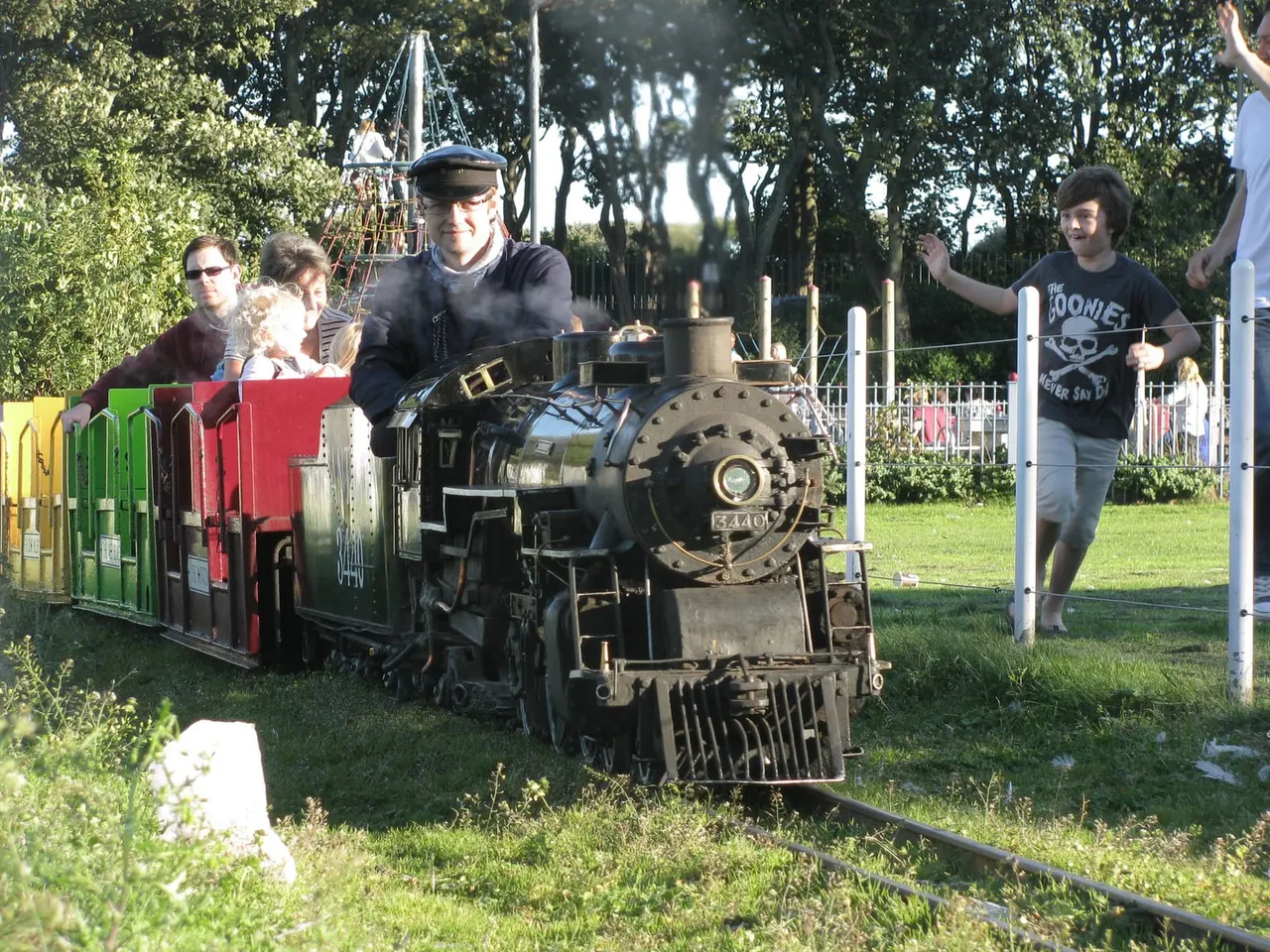 Miniature steam locomotive with passengers riding in colourful carriages through a tree-lined park setting.