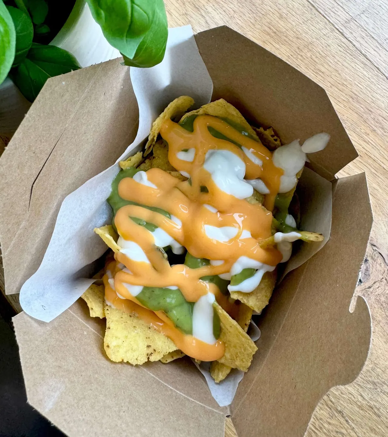 Stacked tortilla chips topped with melted cheese sauce, sliced peppers and soured cream in a cardboard takeaway box.