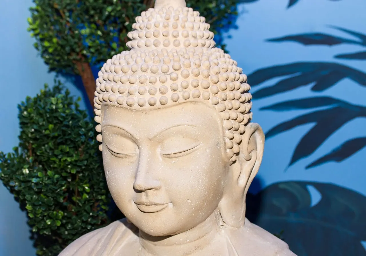 Cream stone Buddha head sculpture with eyes closed, beside a topiary bush and blue mural backdrop.