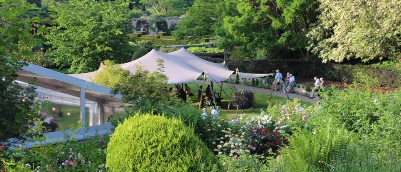 Formal gardens with flowering borders and topiary surrounding a cream canvas marquee, with visitors strolling on the lawn and a modern pavilion visible to the left.