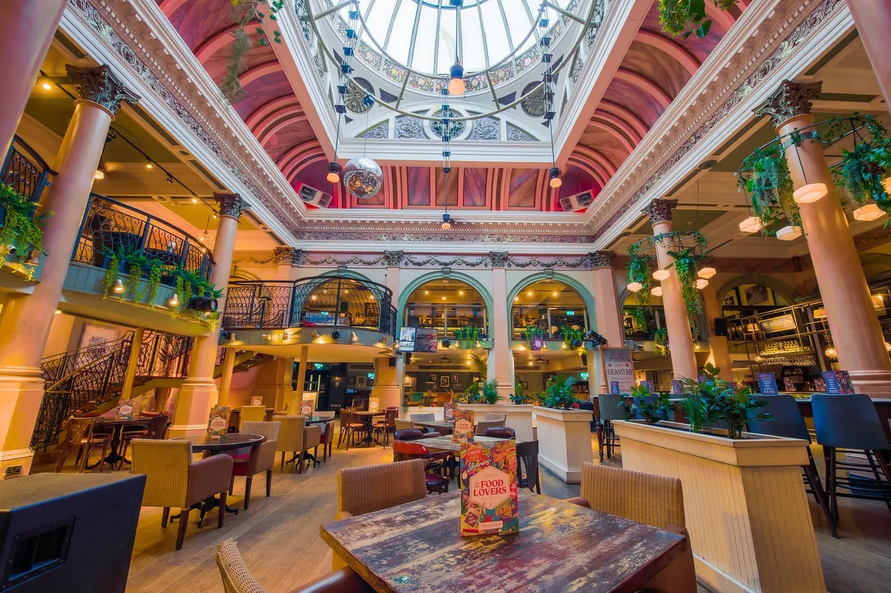 Ornate indoor atrium with glass domed ceiling, multiple levels with balconies, decorative columns, potted plants and warm lighting throughout the space.