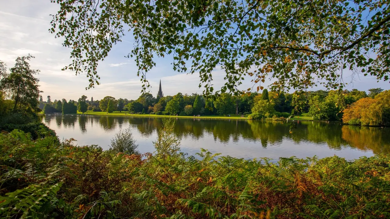 River with tree-lined banks and a distant church spire, viewed through overhanging foliage in autumn.