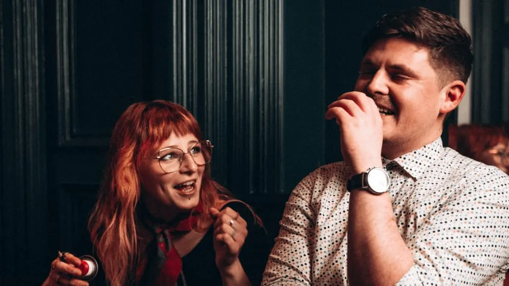 Two people singing together at a karaoke event against a dark corrugated metal backdrop.