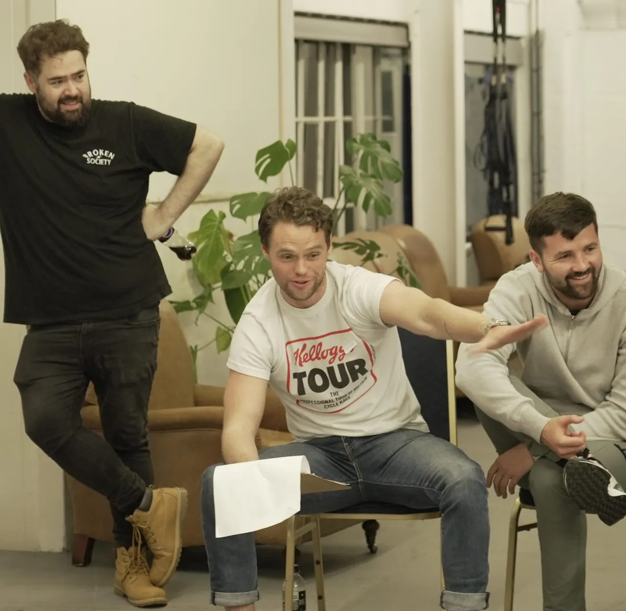 Three men having fun in an indoor leisure space, seated and standing around a sofa with potted plants nearby.