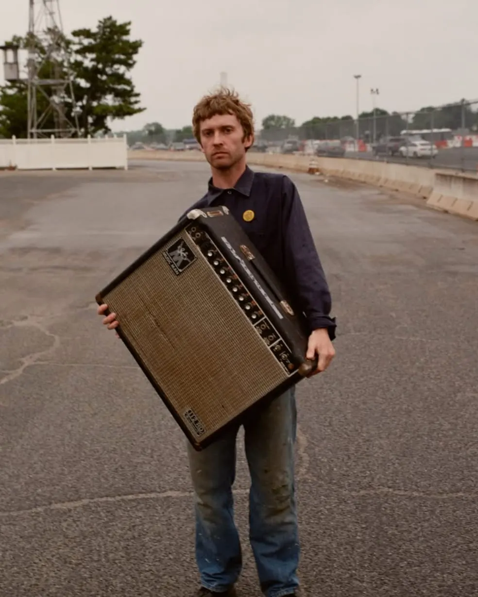 Man holding a guitar amplifier on an empty carpark with industrial buildings in the background.
