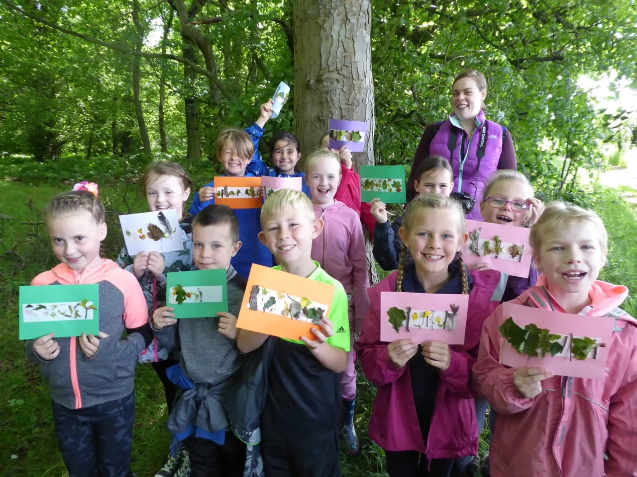 Group of children and an adult holding pressed plant displays in a wooded forest setting.