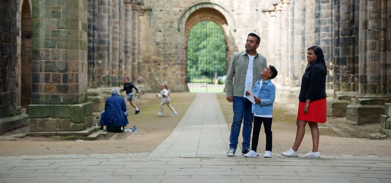 Family visiting the ruins of a medieval abbey, with stone archways and other visitors playing in the background.