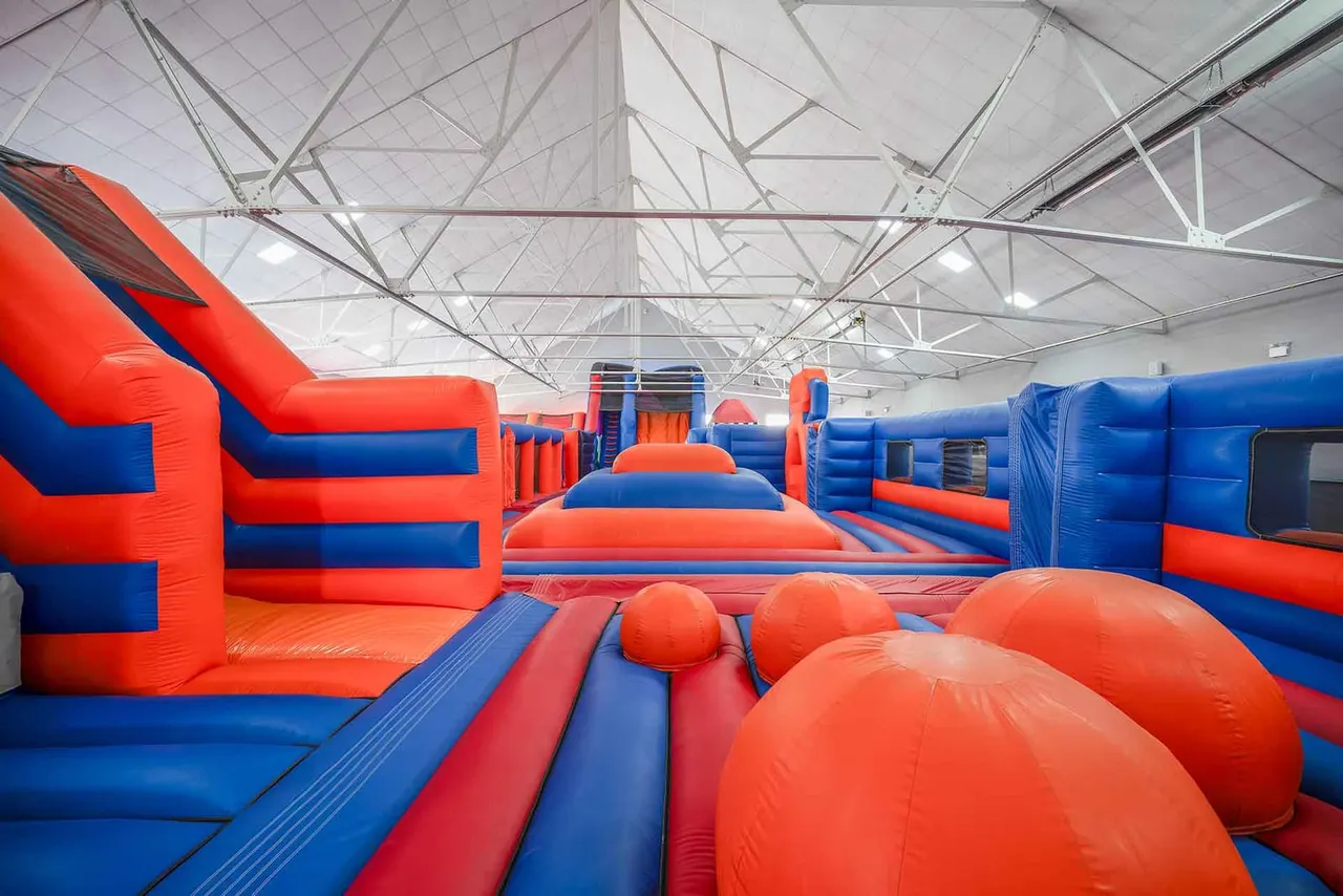 Indoor soft play facility with multiple inflatable structures in red and blue, including slides, obstacles and spheres under a high industrial ceiling.