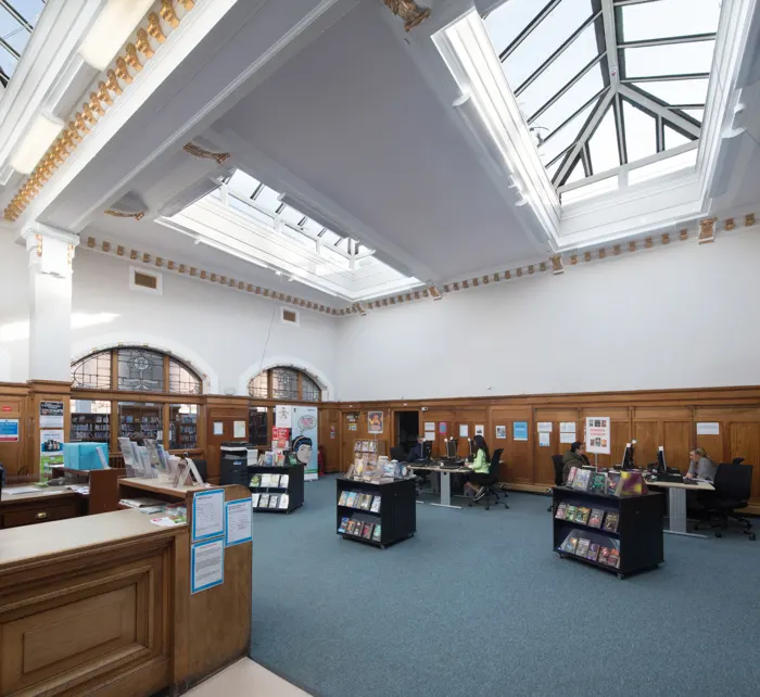 Interior of a library with wooden counters, display stands, and skylights, with staff and visitors inside.