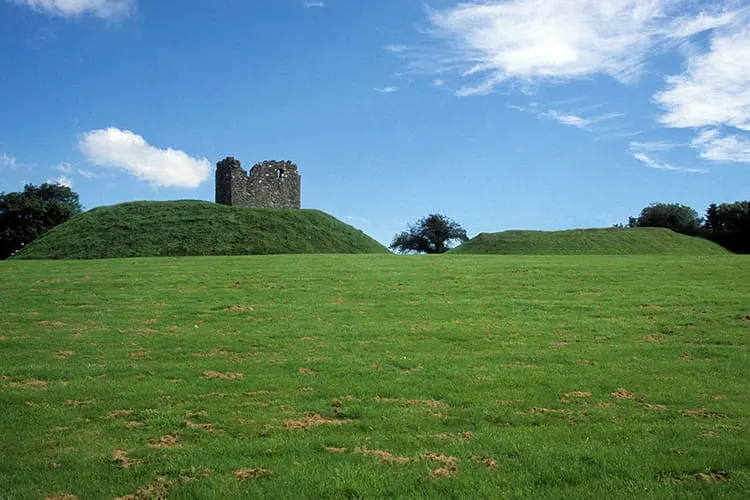 Ruined stone tower standing on top of a grass-covered mound beneath blue sky.