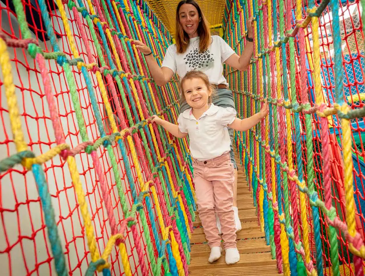 A woman and young child navigate a colourful rope net tunnel during an indoor play activity.